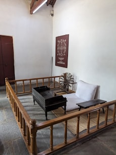 A simple, traditional room with minimal furniture, featuring a low wooden bed and desk, a spinning wheel, and a wall poster with text and a circular design. The space is enclosed by a wooden railing and has a closed wooden door on one side.