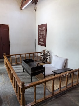 A simple, traditional room with minimal furniture, featuring a low wooden bed and desk, a spinning wheel, and a wall poster with text and a circular design. The space is enclosed by a wooden railing and has a closed wooden door on one side.