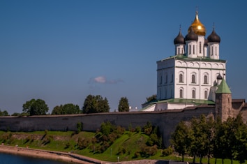A white Orthodox church with green rooftops and gilded domes sits atop a hill surrounded by a stone wall. The building has multiple levels and is adorned with several arches and intricately designed windows. The foreground features a green landscape with grass, shrubs, and trees. A river is visible in the bottom left corner of the image under a clear blue sky.