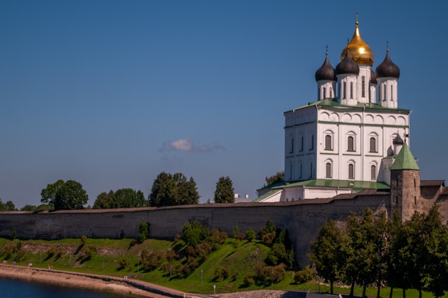 A white Orthodox church with green rooftops and gilded domes sits atop a hill surrounded by a stone wall. The building has multiple levels and is adorned with several arches and intricately designed windows. The foreground features a green landscape with grass, shrubs, and trees. A river is visible in the bottom left corner of the image under a clear blue sky.