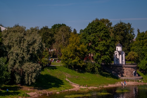A tranquil park landscape features lush green trees and a small, white chapel-like structure with an arched entrance and a dark dome. A stone wall leads down to a grassy area beside a calm body of water where a few people are gathered near the shore.