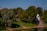 A tranquil park landscape features lush green trees and a small, white chapel-like structure with an arched entrance and a dark dome. A stone wall leads down to a grassy area beside a calm body of water where a few people are gathered near the shore.