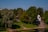 A tranquil park landscape features lush green trees and a small, white chapel-like structure with an arched entrance and a dark dome. A stone wall leads down to a grassy area beside a calm body of water where a few people are gathered near the shore.