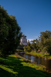 A serene landscape featuring a historic building with golden domes, partially obscured by lush trees. A tranquil river flows in the foreground, reflecting the greenery and blue sky. The setting suggests a peaceful, natural environment.