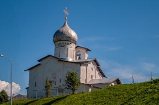 An old, white Orthodox church with a distinctive silver onion dome sits atop a green hill. A cross adorns the top of the dome, and the building's walls show signs of age and wear. The sky is clear and blue, providing a striking contrast to the church's light-colored structure. A few small trees are scattered in front of the building, and a lamppost is visible on the left side.