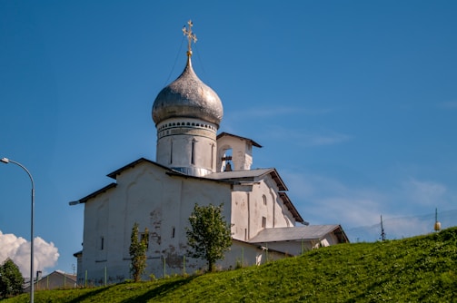 An old, white Orthodox church with a distinctive silver onion dome sits atop a green hill. A cross adorns the top of the dome, and the building's walls show signs of age and wear. The sky is clear and blue, providing a striking contrast to the church's light-colored structure. A few small trees are scattered in front of the building, and a lamppost is visible on the left side.