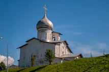 An old, white Orthodox church with a distinctive silver onion dome sits atop a green hill. A cross adorns the top of the dome, and the building's walls show signs of age and wear. The sky is clear and blue, providing a striking contrast to the church's light-colored structure. A few small trees are scattered in front of the building, and a lamppost is visible on the left side.