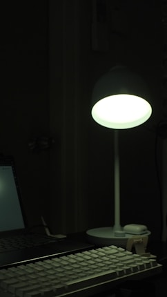 A dimly lit workspace featuring a glowing desk lamp illuminating a keyboard and part of a laptop. The light creates a soft ambiance in the otherwise dark room.