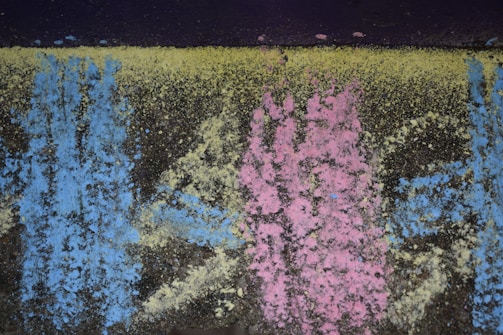 Colorful chalk powder patterns of blue, pink, and yellow cover a dark, rough textured surface. The colors are dispersed unevenly, creating an abstract and organic appearance.