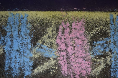 Colorful chalk powder patterns of blue, pink, and yellow cover a dark, rough textured surface. The colors are dispersed unevenly, creating an abstract and organic appearance.