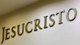 The image features large, bold letters spelling out the word 'JESUCRISTO' on a plain wall. The letters are three-dimensional with a metallic or bronze appearance, casting shadows that add depth.