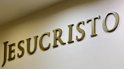 The image features large, bold letters spelling out the word 'JESUCRISTO' on a plain wall. The letters are three-dimensional with a metallic or bronze appearance, casting shadows that add depth.