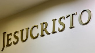 The image features large, bold letters spelling out the word 'JESUCRISTO' on a plain wall. The letters are three-dimensional with a metallic or bronze appearance, casting shadows that add depth.