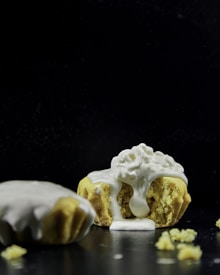 A partially crumbled pastry topped with a generous amount of creamy white icing flows over the sides, surrounded by a few scattered crumbs. The background is dark, which highlights the texture and color of the pastry and icing.