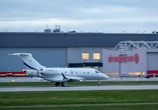 An airplane with Canadian markings on its tail is taxiing on a runway at an airport. The aircraft is white with blue accents and has the registration number C-GASK. In the background, there is a large building with the Air Canada logo visible, suggesting the location is a commercial airport.