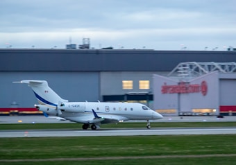 An airplane with Canadian markings on its tail is taxiing on a runway at an airport. The aircraft is white with blue accents and has the registration number C-GASK. In the background, there is a large building with the Air Canada logo visible, suggesting the location is a commercial airport.