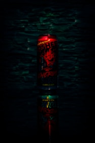 A red energy drink can with bold, fiery graphics and text, set against a dark textured background. The can is reflected below, creating a mirrored effect.