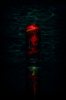 A red energy drink can with bold, fiery graphics and text, set against a dark textured background. The can is reflected below, creating a mirrored effect.