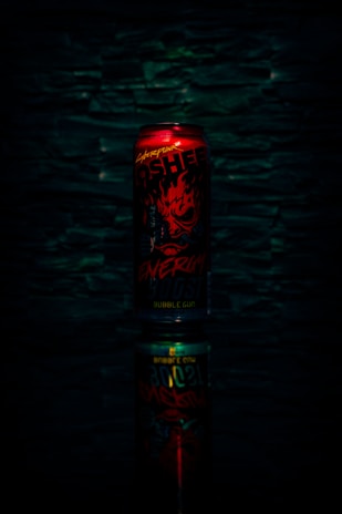 A red energy drink can with bold, fiery graphics and text, set against a dark textured background. The can is reflected below, creating a mirrored effect.