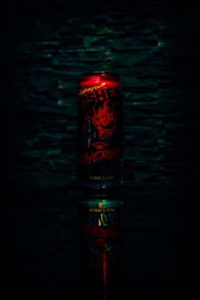 A red energy drink can with bold, fiery graphics and text, set against a dark textured background. The can is reflected below, creating a mirrored effect.