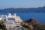 White buildings with domed rooftops are perched on a hillside overlooking a deep blue sea. The architecture is classic of the Greek islands, with whitewashed walls and blue accents. In the background, a mountainous landscape stretches across the horizon under a clear sky.