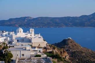 White buildings with domed rooftops are perched on a hillside overlooking a deep blue sea. The architecture is classic of the Greek islands, with whitewashed walls and blue accents. In the background, a mountainous landscape stretches across the horizon under a clear sky.