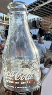 A close-up view of a glass Coca-Cola bottle with the logo and text 'Sabor Original' in the foreground, with a blurred background of an outdoor setting where people are seated and a small stage with a musician is visible.