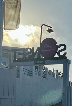 A sign with bold letters is silhouetted against a bright sky with the sun partially obscured by clouds. There is a banner with text and a lamp post near the sign. The setting suggests a warm, possibly coastal location.