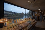 A shop interior with a large window offering a view of a calm river and traditional houses with gabled roofs outside. The shop displays numerous small figurines and goods on wooden tables. Several lamps provide soft lighting, creating a cozy atmosphere. A person browses through the items on the right side.
