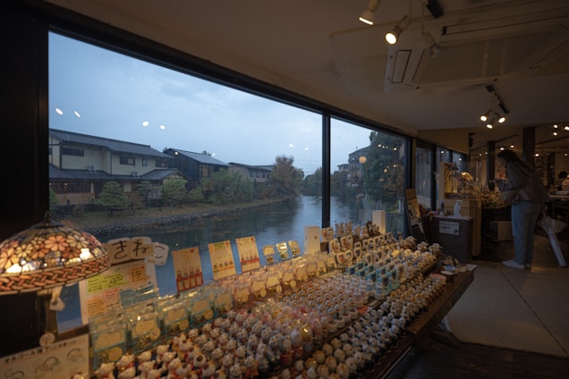 A shop interior with a large window offering a view of a calm river and traditional houses with gabled roofs outside. The shop displays numerous small figurines and goods on wooden tables. Several lamps provide soft lighting, creating a cozy atmosphere. A person browses through the items on the right side.