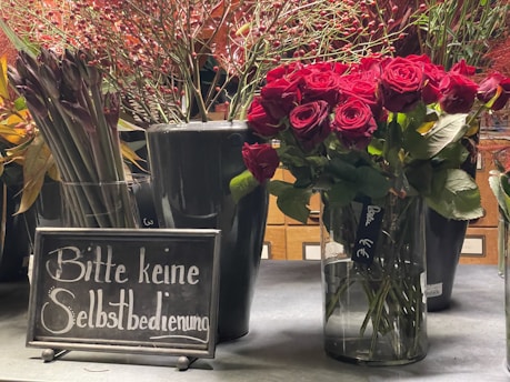 Several vases filled with different types of flowers, including a large bouquet of vivid red roses, are displayed on a counter. Next to the vases, a sign in German reads 'Bitte keine Selbstbedienung,' which translates to 'Please, no self-service.'