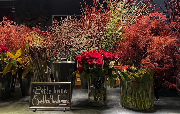 A collection of vibrant floral arrangements, primarily featuring red flowers and green foliage. The flowers are placed in glass vases on a flat surface, and there is a sign with German text in front of them. The backdrop is dark, accentuating the rich colors of the flowers.
