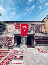 A historic stone building adorned with a variety of colorful, intricate carpets and rugs displayed on both the walls and the ground. The centerpiece is a large Turkish flag hanging prominently in the middle, adding a sense of national pride. The scene is set under a clear blue sky, contributing to a vibrant and lively atmosphere.
