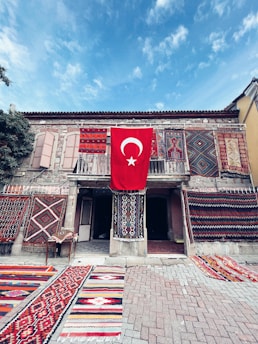 A historic stone building adorned with a variety of colorful, intricate carpets and rugs displayed on both the walls and the ground. The centerpiece is a large Turkish flag hanging prominently in the middle, adding a sense of national pride. The scene is set under a clear blue sky, contributing to a vibrant and lively atmosphere.