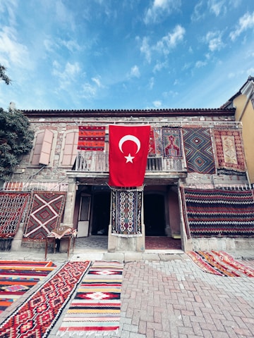 A historic stone building adorned with a variety of colorful, intricate carpets and rugs displayed on both the walls and the ground. The centerpiece is a large Turkish flag hanging prominently in the middle, adding a sense of national pride. The scene is set under a clear blue sky, contributing to a vibrant and lively atmosphere.