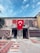 A historic stone building adorned with a variety of colorful, intricate carpets and rugs displayed on both the walls and the ground. The centerpiece is a large Turkish flag hanging prominently in the middle, adding a sense of national pride. The scene is set under a clear blue sky, contributing to a vibrant and lively atmosphere.