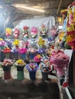 A vibrant display of flower arrangements in various colors and styles is presented on shelves. The scene includes an array of bouquets in different colored wrappings, with a backdrop of a light-colored cloth and some rustic elements like wooden beams and foliage.