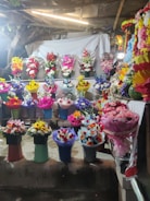 A vibrant display of flower arrangements in various colors and styles is presented on shelves. The scene includes an array of bouquets in different colored wrappings, with a backdrop of a light-colored cloth and some rustic elements like wooden beams and foliage.