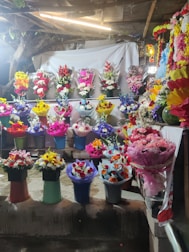 A vibrant display of flower arrangements in various colors and styles is presented on shelves. The scene includes an array of bouquets in different colored wrappings, with a backdrop of a light-colored cloth and some rustic elements like wooden beams and foliage.