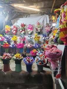 A vibrant display of flower arrangements in various colors and styles is presented on shelves. The scene includes an array of bouquets in different colored wrappings, with a backdrop of a light-colored cloth and some rustic elements like wooden beams and foliage.