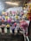 A vibrant display of flower arrangements in various colors and styles is presented on shelves. The scene includes an array of bouquets in different colored wrappings, with a backdrop of a light-colored cloth and some rustic elements like wooden beams and foliage.