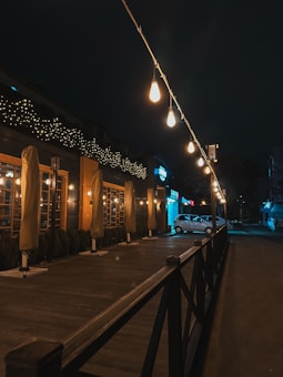 A dimly lit outdoor area featuring a series of hanging light bulbs strung across the dark sky. Below, a wooden platform with closed patio umbrellas runs alongside a building adorned with fairy lights. A white car is parked nearby, and the setting gives off a cozy, tranquil atmosphere.