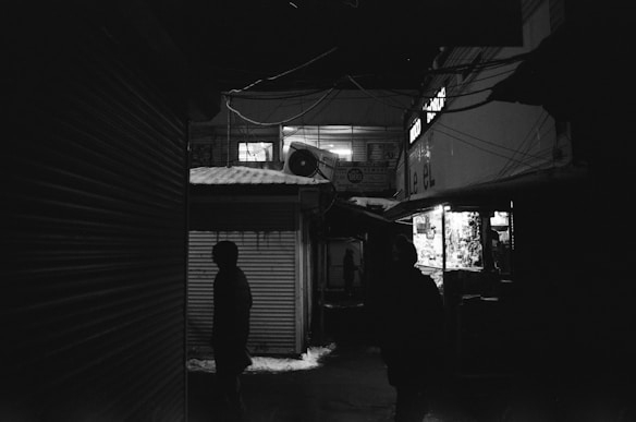 Dark alleyway scene with silhouettes of people walking past dimly lit shops. Overhead wires crisscross between buildings, and there is a visible air conditioning unit on the wall. The atmosphere is moody and shadowy with few light sources coming from windows and signage.