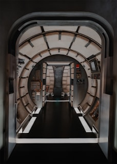 A futuristic, metallic corridor with a round, tunnel-like design. The structure features pipes and consoles, giving a spaceship or space station vibe. Ambient lighting illuminates the corridor, highlighting the sleek, industrial look.