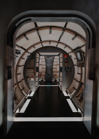 A futuristic, metallic corridor with a round, tunnel-like design. The structure features pipes and consoles, giving a spaceship or space station vibe. Ambient lighting illuminates the corridor, highlighting the sleek, industrial look.
