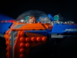A close-up of a detailed LEGO spaceship model, featuring a minifigure inside a transparent dome cockpit. The model showcases vibrant colors with orange and blue bricks, as well as intricate design elements and small green accents.
