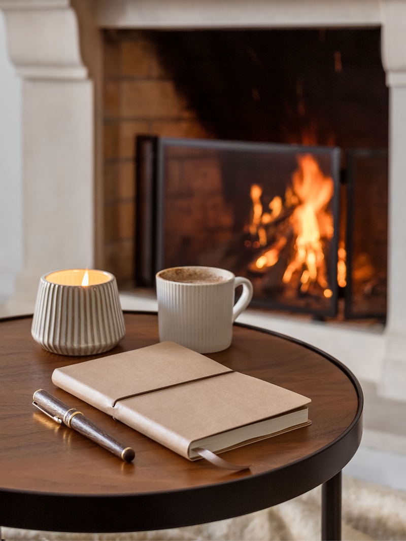 A notebook and pen on a table beside a candle and a mug, with a fireplace softly blurred in the background