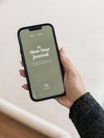 A smartphone displaying a digital journal cover titled 'The New Year Journal' for the years 2023-2024 is being held in a person's hand. The cover has a minimalist design with a greenish background and white text, and the word 'PROPHESEE' at the bottom. The person's hand is visible, showing manicured nails, and they are wearing a dark sweater.