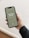 A smartphone displaying a digital journal cover titled 'The New Year Journal' for the years 2023-2024 is being held in a person's hand. The cover has a minimalist design with a greenish background and white text, and the word 'PROPHESEE' at the bottom. The person's hand is visible, showing manicured nails, and they are wearing a dark sweater.