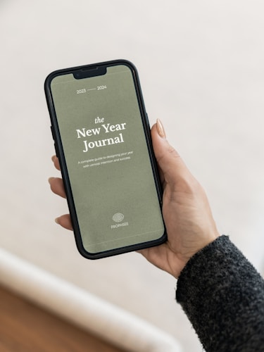 A smartphone displaying a digital journal cover titled 'The New Year Journal' for the years 2023-2024 is being held in a person's hand. The cover has a minimalist design with a greenish background and white text, and the word 'PROPHESEE' at the bottom. The person's hand is visible, showing manicured nails, and they are wearing a dark sweater.